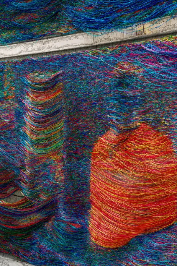 Photo of colorful threads—mostly blue and orange—woven around what appears to be a horizontal bar. Below, a cylindrical shape and another shape resembling a bust protrude.